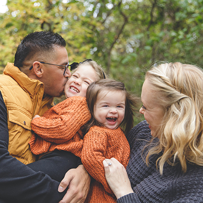 a close up family photo of dad, two young girls in orange, and mom in gray, the girls both have large smiles