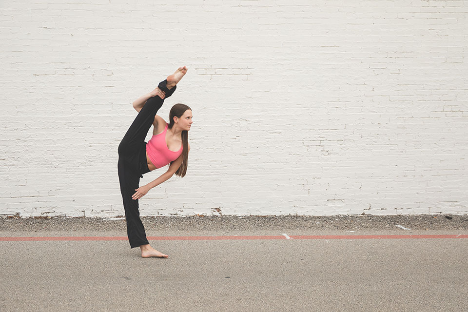 light skinned teenage girl in a pink top and black pants in an elevated leg dance pose in front of a white brick wall - dallas dance photography