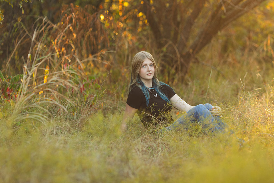 photo of a senior girl with light skin sitting in tall grass with warm natural tones behind her - plano senior photography