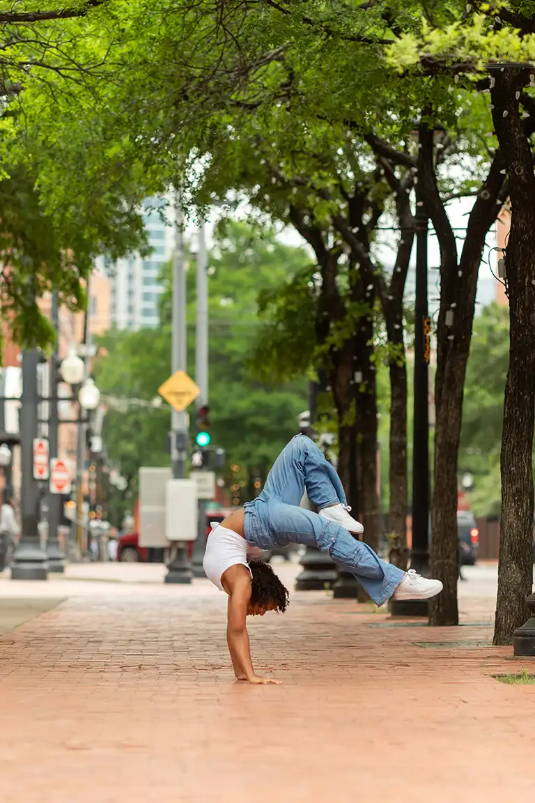 vertical image of a brick sidewalk with trees and a Black teenage girl in white shirt and jeans balancing on her hands with her legs over - dallas dance photography