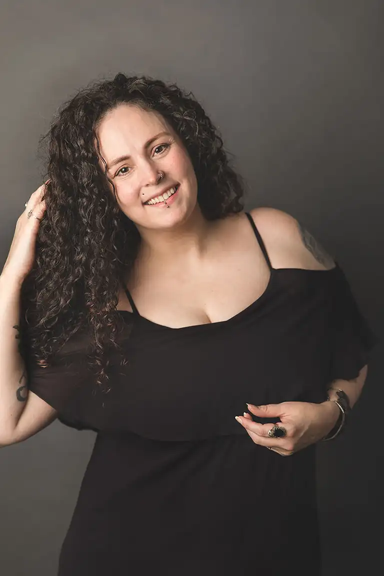 light skinned woman with dark curly hair and a black dress on a dark gray studio background - I am smiling at the camera with one hand at my hair - plano photographer brandi thompson