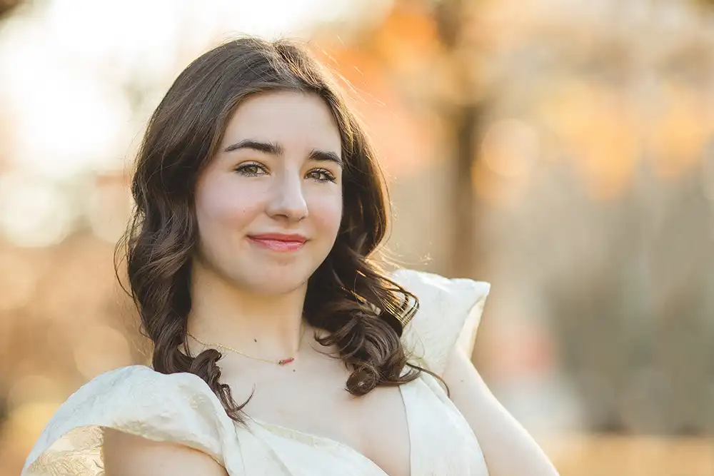 horizontal headshot of light skinned teenage girl with dark hair wearing a cream dress, and a blurred warm background behind her - plano senior photography